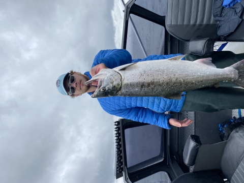 Angler in blue jacket, with hat and sunglasses, stands on a boat holding a large Chinook salmon with the Columbia River in the background