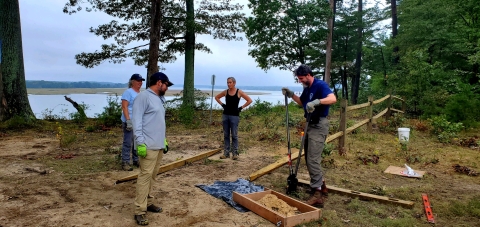 four people work to build a fence surrounded by trees and a river in the distance