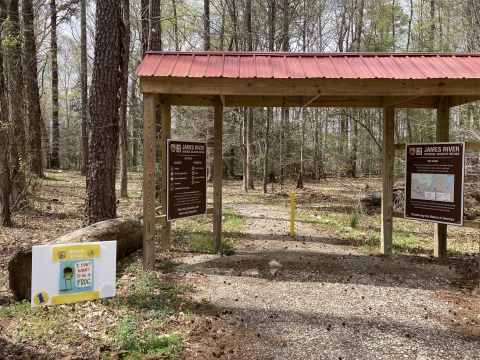 Entrance to the Powell Creek Trail sheltered by a wooden archway pavilion and two kiosk signs: one with the James River NWR trail map and the other with prohibited items/actions. The first story trail sign is located in front of the pavilion with the cover of "I Don't Want to be a Frog" pictured.