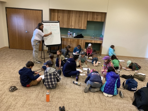 A Ranger pointing to a white board, students sitting on the ground working on project.