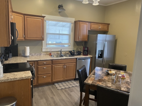 Picture taken from a doorway. An L-shaped countertop wraps around the wall. Grey floorboards and a small table with black chairs fill the middle of the room.