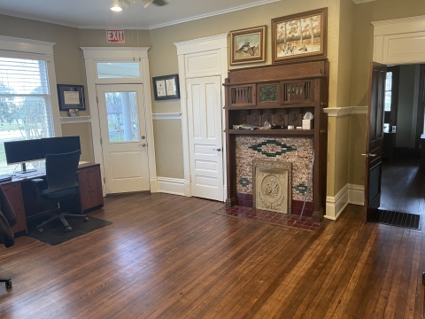 Dark wooden floor boards reflect light coming through the windows. A desk with two monitors and office chair are placed against the front window. Two white doors with white trim wrap around the corner. A dark wooden mantle and cabinet enclose a fireplace. Two duck paintings hang high on the wall. An open door reveals a peak into an adjacent dark room.