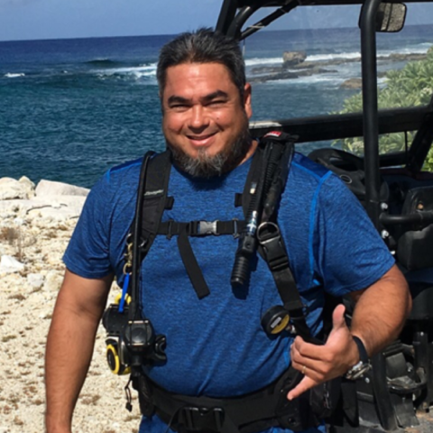 Matthew Onellion gives a shaka to the camera. He is wearing a blue shirt and standing on a beach.