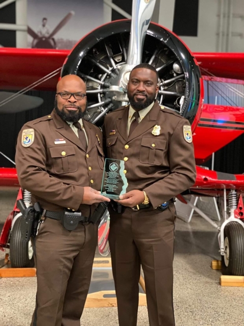 2 African American men stand in front of plane; one holds award