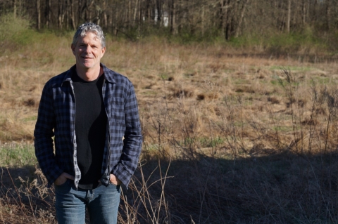 A man in a blue shirt stands in front of a field. He is facing the camera and smiling, with his hands in his pockets. 