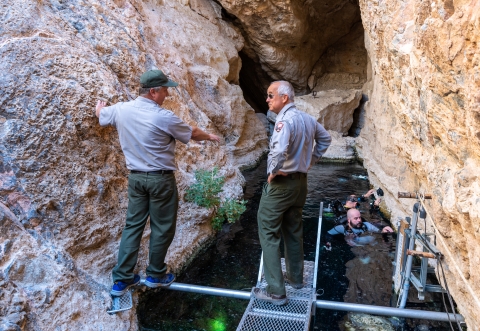 Two uniformed men stand on a platform constructed over a water-filled crevasse while two men in diving gear swim below. 