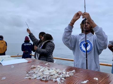 Youth learn to hook bait on fishing rod. Cut up sardines are available in a mound on top of a live bait tank.