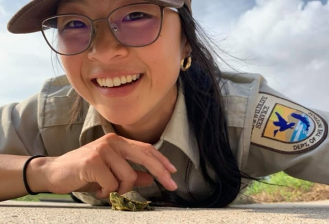 A woman in a U.S. Fish and Wildlife Service uniform smiling as she lies on the ground with a tiny turtle crawling in front of her