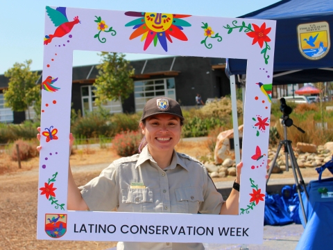 A woman in a brown U.S. Fish and Wildlife Service uniform with a broad smile on her face holding a frame around herself. On the frame are the words Latino Conservation Week.