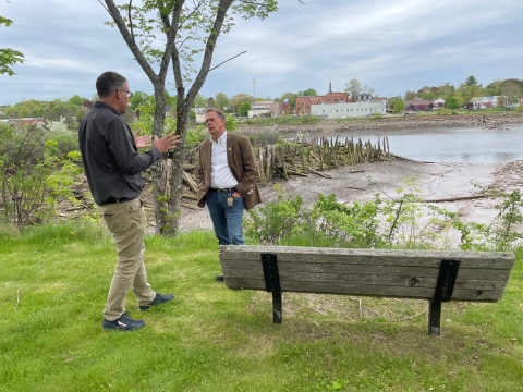 A man in a brown sports jacket stands alongside a river with another gentleman. The two are in active conversation as they stand near a bench. In the background residential and commercial building line the edge of the river. 