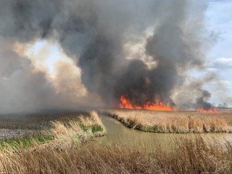 Fire on right and thick dark smoke above fields split with water