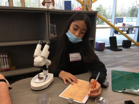 A child writes notes on a paper next to a microscope