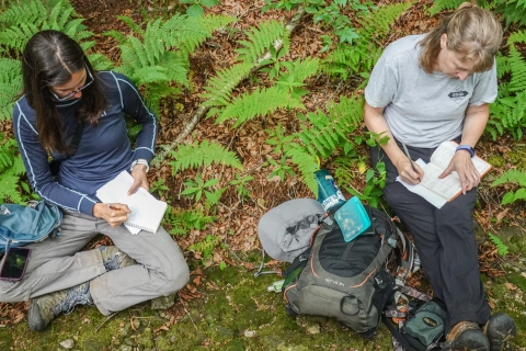 Two people sitting on the ground, each writing in a notebook