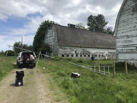 Two people look into the back of a vehicle behind a slender antenna tower lying on its side. Nearby are two large old barns.