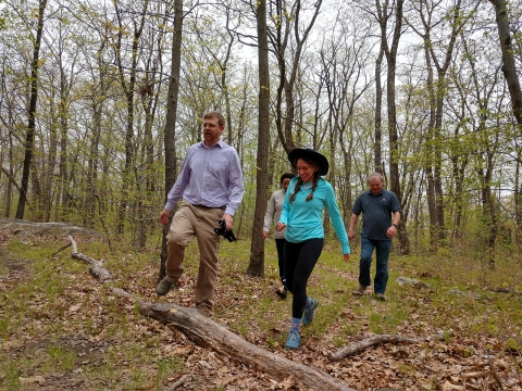 A group of four people walk along a path in the woods during the early summer. They step over a log that lays across the trail. There is low underbrush and brown leaves across the forest floor. 