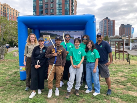 eight people stand in front of an inflatable cube object. The people are smiling, and two of them hold archery bows. They appear to be in an urban park, as tall buildings appear behind them.