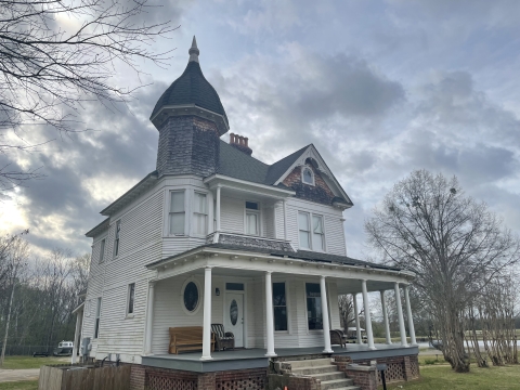 White two-story house with a front porch, nine columns, and a dark roof in the foreground with grey skies behind it.