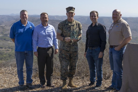 five men pose on hilltop with one in military uniform holding a plaque 