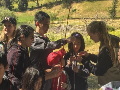 A group of people hold fishing rods while a woman demonstrates a technique.