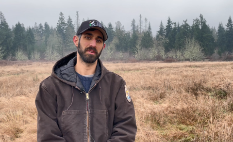 A man in brown jacket a baseball hat stands in a field