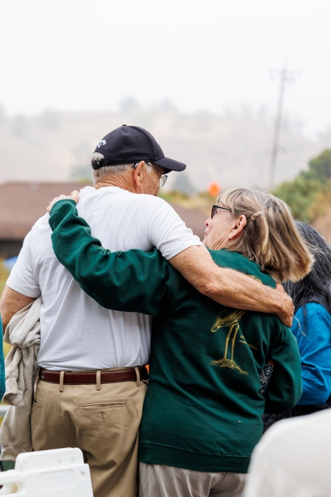 Two people stand arm in arm with their backs turned to the camera. 