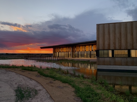 a large building surrounded by a pond. The sun sets behind dramatic clouds in the background