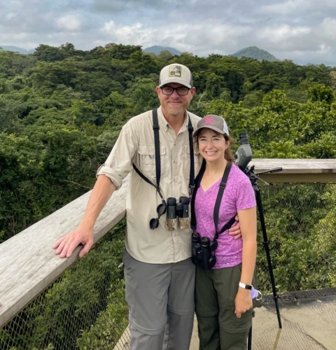 A man and woman smiling together with binoculars around their necks, stand on a high deck overlooking a forest