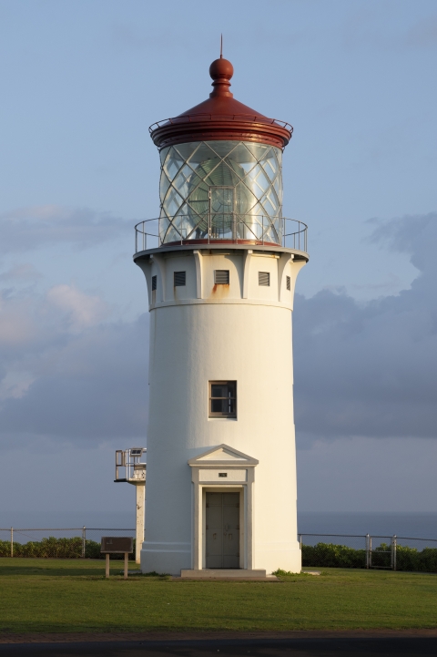 a lighthouse fills the frame. The sky is an early morning blue with wisps of purple. It is partially cloudy. The lighthouse is on a grassy point with a fence and the ocean behind it. 