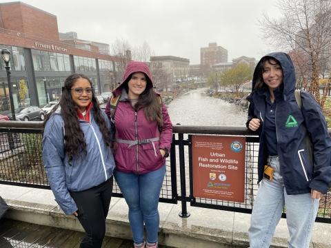 3 young woman stand on a bridge overlooking a city landscape and river.