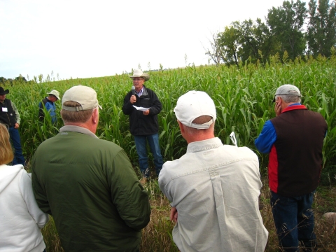 A man wearing a cowboy hat stands in front of a field, speaking to a small crowd of people