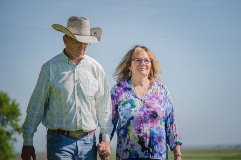 A man wearing a cowboy hat and a woman walk hand-in-hand across a grassy field