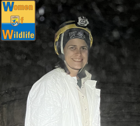 Pam McDill smiles at the camera as she stands in dark cave. She is wearing a white jumpsuit and a helmet with a headlamp on it. The WoW: Women of Wildlife logo is in the upper left corner of the image.