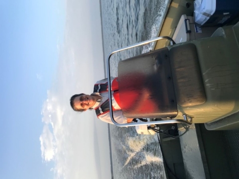 A woman in an orange life vest smiles at the helm of a boat