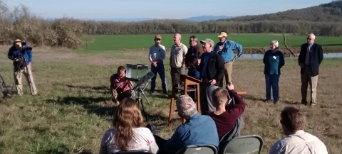 A man speaks at a podium holding a microphone with spectators and cameras around him in a grassy area with mountains in the background