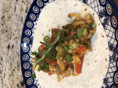 A colorful plate with a tortilla filled with fish fajita ingredients and topped with a sprig of rosemary for garnish.