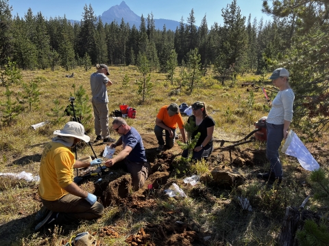 Six people hold tools around a hole in an alpine meadow