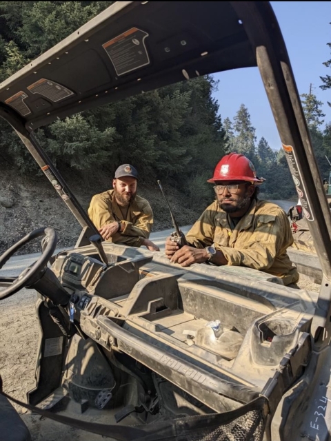 2 people in firefighting gear lean on hood of vehicle