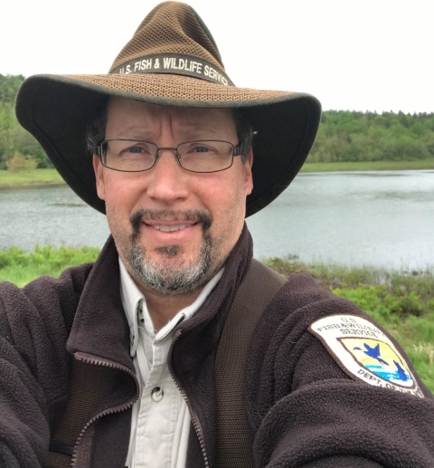 A person in a brown U.S. Fish and Wildlife Service uniform and hat, stands in front of a pond surrounded by green trees