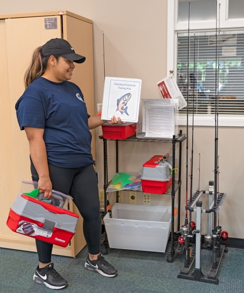 Service intern holding fishing tackle box and a booklet inside of a library next to fishing rods and more tackle boxes. 