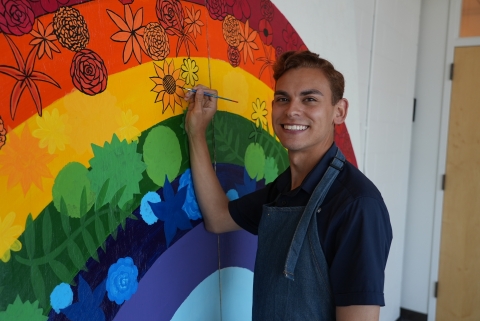 a man smiles while painting a large rainbow mural decorated with various flowers