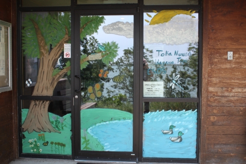 Artwork painted onto the front doors of a visitor center. The artwork depicts a tree, a pond with ducks, butterflies, and a sun behind a cloud. Words painted read Toka Nuwan Wayawa Tipi ESDS