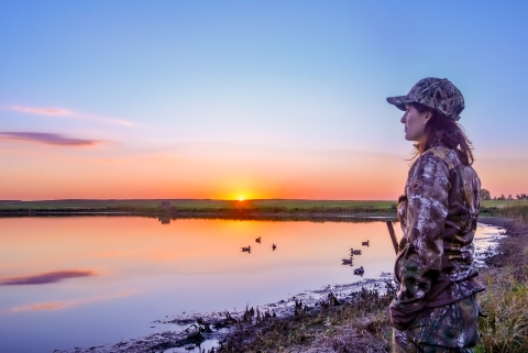 A woman waterfowl hunting by a wetland at sunset