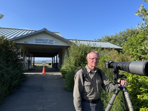 Person standing in front of a building with their hand on a tripod that is holding a camera with large lens