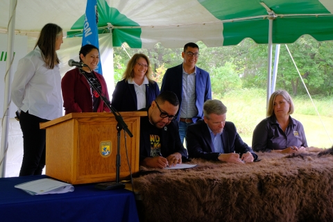 Four people stand behind three people sitting at a table. Two men seated at the table are signing documents in front of them. A brown fur is draped over the table