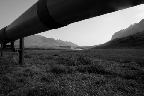 A black and white photo of a section of the Trans-Alaska Oil Pipeline in the foreground -- underneath it, permafrost tundra stretches across vast hills. In the background, peaks of the Brooks Range, within the Arctic National Wildlife Refuge, extend into a white sky.