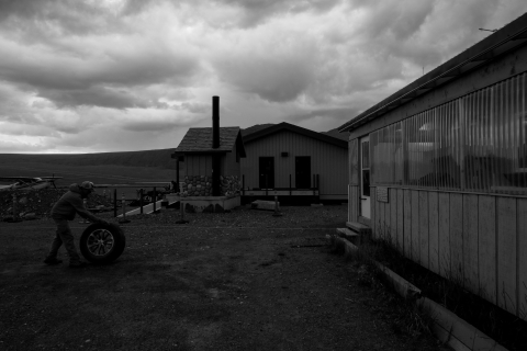 A black and white photo -- in the foreground, on the right, a profile of the rectangular USFWS bunkhouse stretches into the background, where a new, nearly-completed bunkhouse stands, centered in the frame. On the left, Jimmy Fox, the Refuge Manager of the Yukon National Wildlife Refuge rolls a spare tire along the gravel. Storm clouds hover above it all.