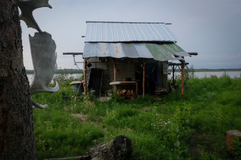 A color photo of Chief Rhonda Pitka's smokehouse, which has been empty for four years due to a Chinook salmon shortage on the Yukon River, which flows just a few feet away. A slanted metal sheet roof extends over a wooden house frame as tall grass grows around it.