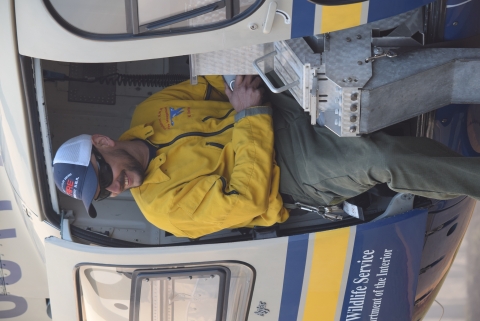 A firefighter sits on the edge of a helicopter, half inside and half out the door.