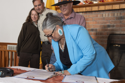 A woman wearing blue signs a document as three people watch from behind her