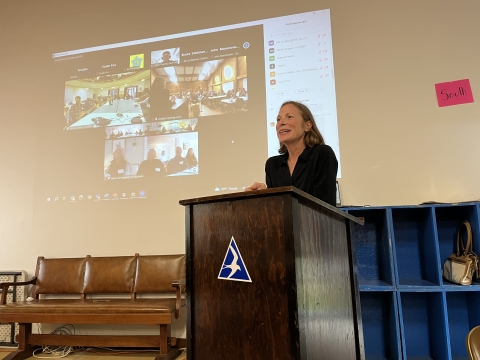 a woman stands at a lectern in front of a screen showing virtual meeting participants